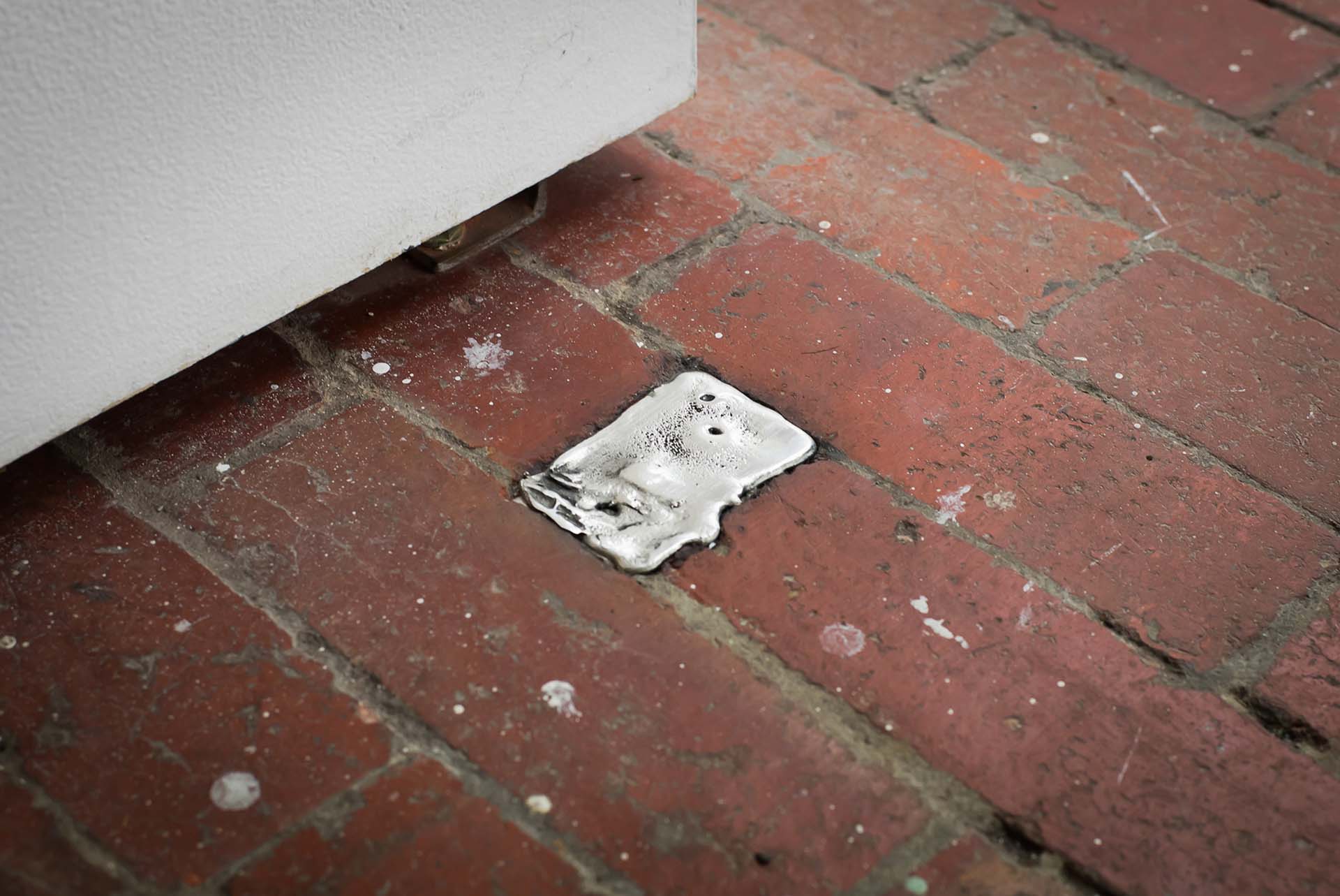 Small silver pewter ingot embedded in a brick floor beneath a white wall